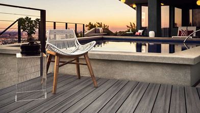 A contemporary rooftop pool area features a woven chair and a small side table with a potted plant. The setting overlooks a cityscape at sunset, creating a relaxed and stylish atmosphere. Warm lighting from the building and the clean lines of the deck enhance the modern aesthetic. No visible text or numbers are present in the image.