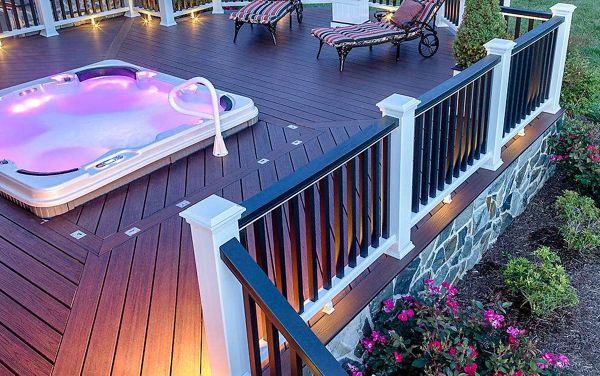 A contemporary outdoor deck features a built-in hot tub with glowing purple lights, surrounded by stylish railings and stone accents. Two cushioned lounge chairs are positioned nearby, inviting relaxation in this landscaped garden setting. The scene is enhanced by evening lighting and blooming flowers, creating a tranquil and inviting atmosphere.