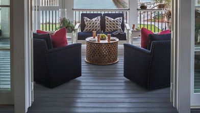 A stylish porch features four dark cushioned armchairs arranged around a round wooden table. The setting overlooks a body of water, visible through the railing, creating a serene and inviting atmosphere. Decorative pillows in navy and red add a pop of color, while copper mugs and small plants enhance the cozy, modern vibe. The scene is well-lit with natural daylight, emphasizing the relaxed, coastal mood.