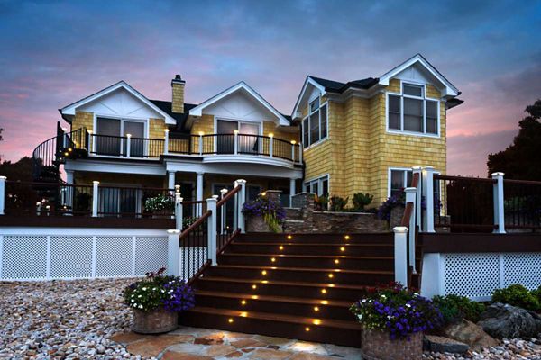 A contemporary two-story home features a prominent staircase with built-in step lighting leading up to a spacious porch. The house exterior combines yellow siding with white trim and large windows, set against a dramatic evening sky. Decorative potted plants and a landscaped stone yard enhance the welcoming atmosphere.