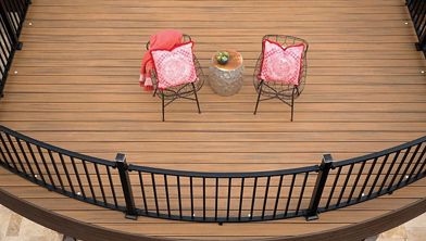 An overhead view shows a curved balcony deck with two wire-frame chairs, each featuring a pink patterned cushion. A small round table with a plant sits between the chairs, and a red throw is draped over one chair. The deck is bordered by a black metal railing and features warm wood tones.