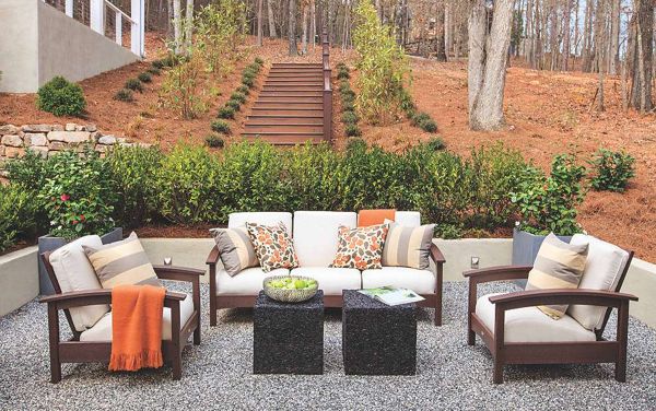 A contemporary outdoor patio features cushioned seating with a sofa and armchairs arranged around two square tables. The setting is in a landscaped garden with gravel flooring, lush greenery, and a staircase leading up a hill. Decorative pillows and a throw blanket add comfort and style, while the neutral and orange color palette complements the natural surroundings.