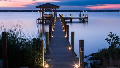 A wooden dock extends over calm water, illuminated by small lights along its edges. The dock leads to a covered boat slip with a boat visible, set against a serene lake at sunset. The sky features soft pink, purple, and blue hues, and the scene is peaceful with no people present.