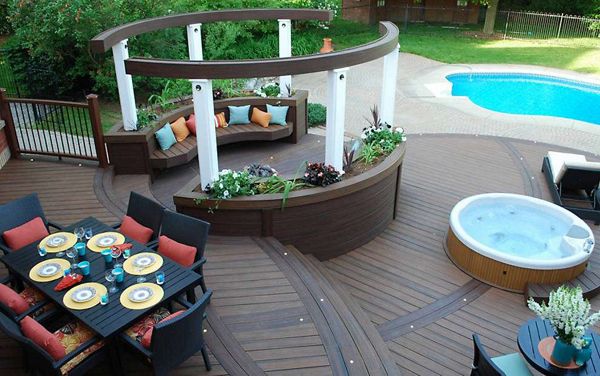A spacious wooden deck features a circular seating area with colorful cushions, a dining table set for eight, and a built-in hot tub. The deck overlooks a bright blue swimming pool and is surrounded by lush greenery. Decorative plants and curved architectural elements add a contemporary touch to the outdoor setting.