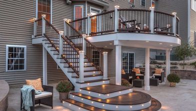 A contemporary two-level outdoor deck features wide, curved steps with built-in LED lighting. The setting is a residential backyard with comfortable seating areas and decorative potted plants. Warm accent lights create a cozy, inviting atmosphere, highlighting the wood and metal railing details. The house exterior is visible in the background, adding to the suburban ambiance.
