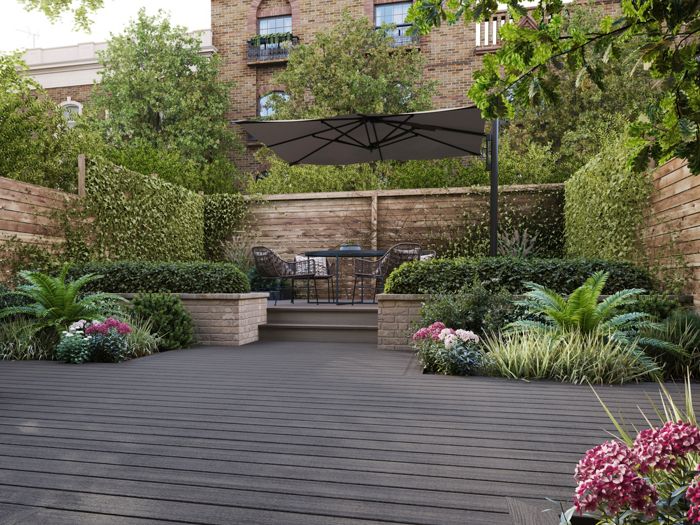 A spacious backyard patio features contemporary outdoor furniture and lush landscaping. The setting includes a raised dining area with a large umbrella, surrounded by wooden fencing and abundant green plants. Decorative flowers and shrubs add color to the scene, while the decked flooring creates a clean, modern look. No visible text or numbers are present in the image.