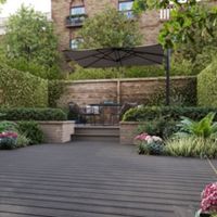 A spacious backyard patio features contemporary outdoor furniture and lush landscaping. The setting includes a raised dining area with a large umbrella, surrounded by wooden fencing and abundant green plants. Decorative flowers and shrubs add color to the scene, while the decked flooring creates a clean, modern look. No visible text or numbers are present in the image.