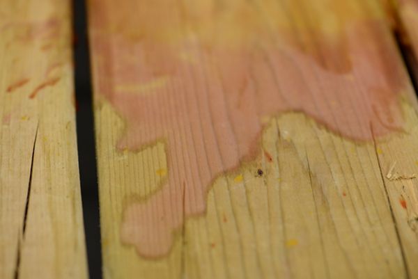 A light-colored wooden surface is shown with a prominent liquid stain spreading across several planks. The setting appears to be indoors, with the focus on the texture and grain of the wood. The stain has an irregular shape and a slightly reddish tint, contrasting with the natural wood color. No visible text, numbers, or identifiable brands are present in the image.
