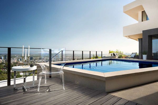 A contemporary rooftop pool sits atop a high-rise building, offering expansive views of the city below. The setting features a sleek glass railing, a small table set for two with candles, and a transparent chair on a wooden deck. The scene is bathed in natural daylight, evoking a serene and luxurious atmosphere. No visible text or numeric values are present in the image.