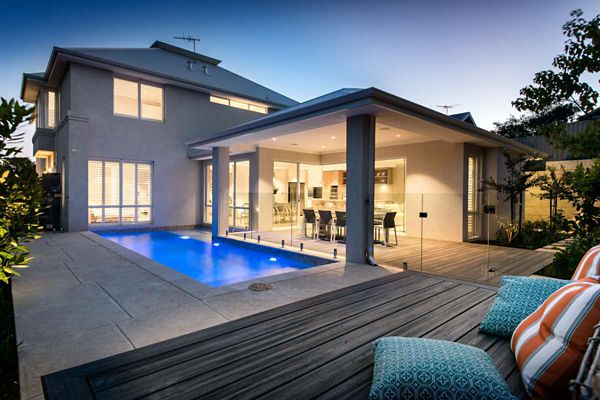A contemporary two-story home features a sleek, illuminated swimming pool adjacent to a covered patio dining area. The setting is at dusk, with warm indoor lights visible through large windows and sliding glass doors. The foreground includes a wooden deck with striped cushions, adding a touch of comfort and color. The overall mood is inviting and tranquil, ideal for evening relaxation or entertaining guests.