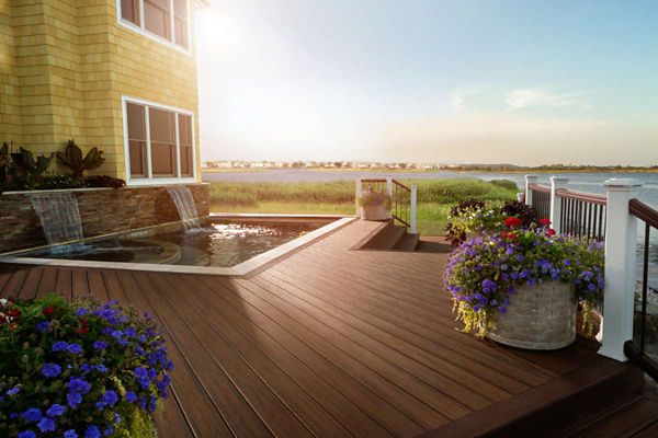 A spacious wooden deck overlooks a serene lake, featuring a built-in hot tub with cascading water features. Large planters filled with vibrant purple and blue flowers add color to the scene. The setting is sunlit, creating a warm and inviting atmosphere, with a yellow house partially visible on the left. The overall mood is tranquil and relaxing, ideal for outdoor leisure.