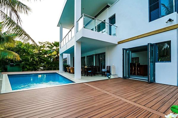 A contemporary two-story house features a large wooden deck adjacent to a rectangular swimming pool. The setting is tropical, with lush palm trees surrounding the outdoor area, and the house has clean white walls and glass balcony railings. Open folding doors connect the indoor and outdoor spaces, creating a seamless flow for relaxation or entertaining. The overall mood is serene and inviting, with natural light enhancing the modern architectural style.