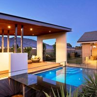 A contemporary backyard features a sleek swimming pool surrounded by glass fencing and a wooden deck. The covered patio area includes built-in seating and a small outdoor kitchen, illuminated by warm recessed lighting. The setting is tranquil, with a clear evening sky and a cozy dining area visible in the background.
