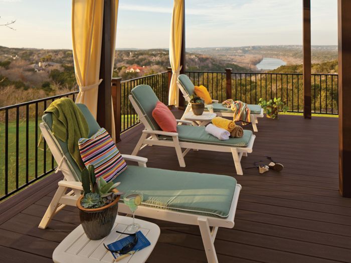 A spacious outdoor deck features several cushioned lounge chairs arranged for relaxation, each with colorful towels and pillows. The setting overlooks a picturesque river valley with rolling hills and distant houses, creating a tranquil atmosphere. A small side table holds a potted cactus, sunglasses, and a drink, while soft evening light enhances the warm, inviting mood. No visible text or numeric values are present in the image.