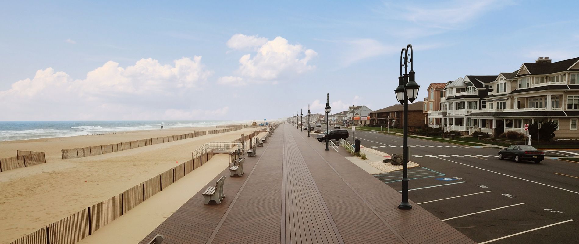 A wide wooden boardwalk stretches along a sandy beach, bordered by a row of benches and classic street lamps. On the right, residential houses line a quiet street with parked cars and visible parking spaces. The scene is calm and inviting, with a clear blue sky and soft clouds overhead, evoking a peaceful coastal atmosphere.