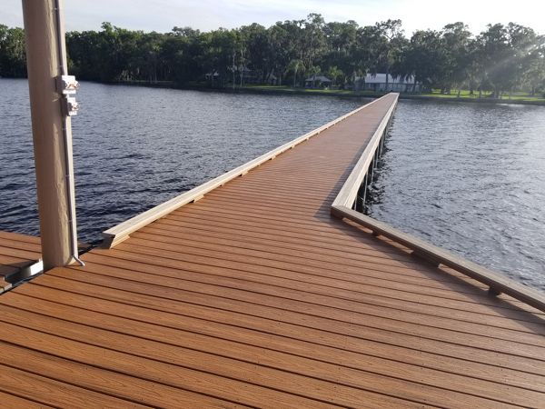 A wide wooden dock stretches out over a tranquil lake, leading toward a distant tree-lined shore. The dock features clean, parallel planks and is bathed in natural daylight, creating a peaceful and inviting atmosphere. The water is calm, reflecting the sky and surrounding greenery, while the far shore is lined with trees and a few visible buildings. No people or text are present in the scene.