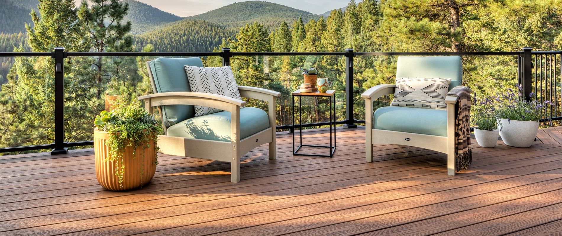 Two cushioned lounge chairs sit on a spacious wooden deck overlooking a lush forest and rolling hills. A small round table between the chairs holds drinks and snacks, while potted plants add color to the scene. The setting is tranquil and inviting, with soft blue and neutral tones creating a relaxing outdoor retreat.