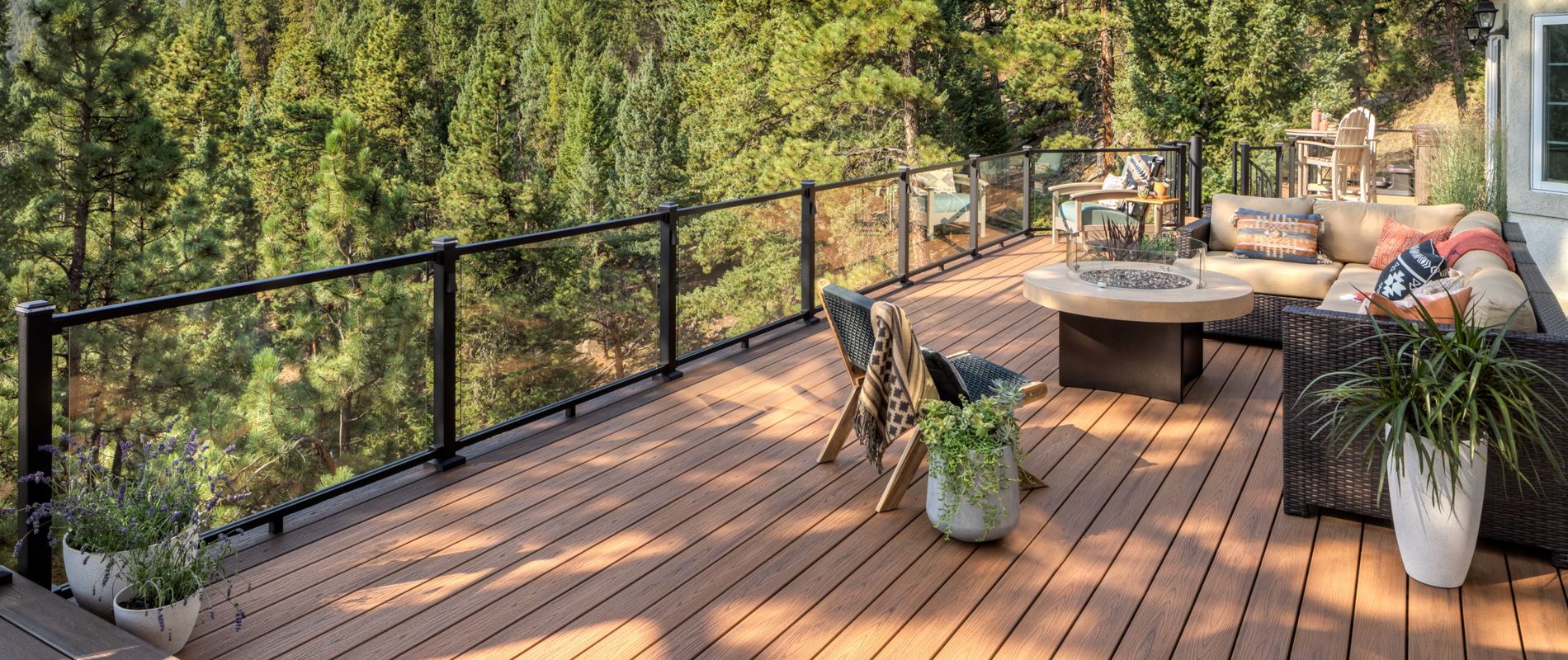 A spacious wooden deck overlooks a dense forest of pine trees, offering a tranquil mountain setting. The deck features contemporary outdoor furniture, a round fire pit table, and decorative potted plants. Glass railing provides an unobstructed view of the natural landscape, while neutral tones and cozy textiles create a relaxing atmosphere. No visible text or numbers are present in the image.