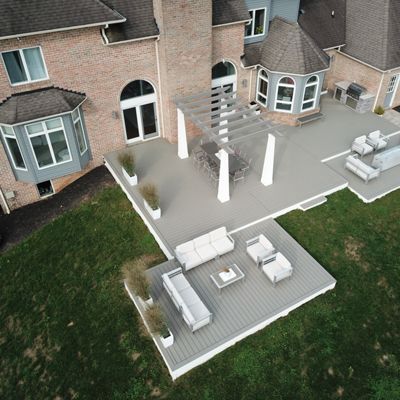 This image shows a spacious backyard patio attached to a brick house, featuring multiple seating areas and a pergola. The deck is furnished with contemporary outdoor sofas, chairs, and a fire pit centerpiece. The setting is suburban, with well-maintained green grass surrounding the patio. The overall mood is inviting and relaxed, with neutral tones dominating the scene. No visible text or numeric values are present in the image.