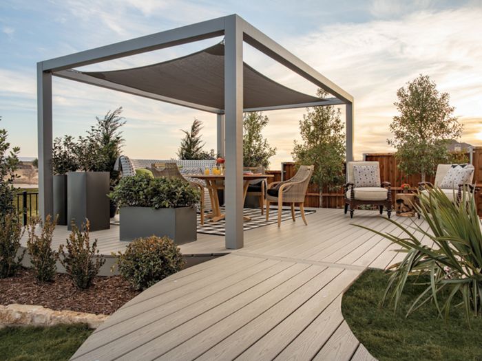A contemporary outdoor patio features a sleek pergola with a fabric canopy, surrounded by lush landscaping and wooden fencing. Comfortable seating and a dining table are arranged under the structure, creating an inviting space for relaxation or gatherings. The setting is enhanced by soft evening light, neutral tones, and well-maintained garden beds. No visible text or numbers are present in the image.