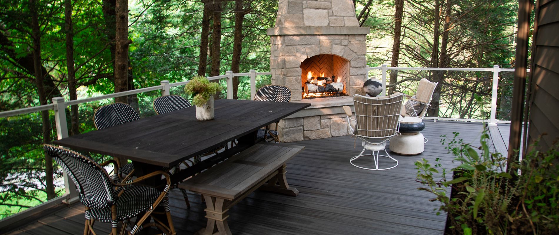 A spacious outdoor deck features a large dark wood dining table with matching chairs and a bench, set amidst a lush green forest backdrop. A stone fireplace with a burning fire creates a warm and inviting atmosphere, while additional seating is arranged nearby for relaxation. The setting is tranquil and stylish, with natural tones and modern outdoor furniture enhancing the cozy ambiance.