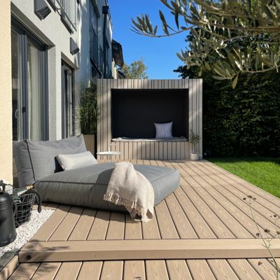 A contemporary outdoor patio features a cushioned sunbed and a stylish cabana with pillows, set against a modern residential building. The wooden deck is bathed in sunlight, surrounded by greenery and a manicured lawn. The scene evokes a relaxed, inviting atmosphere with neutral tones and minimalist design elements.