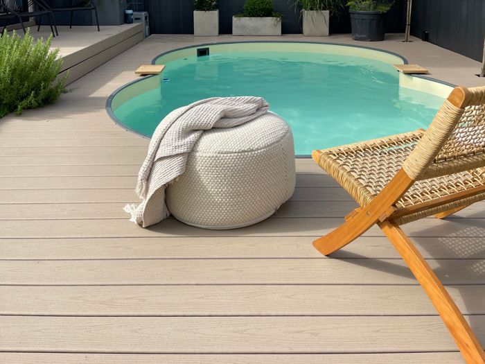 A small, round backyard pool is surrounded by a light wood deck and lush greenery. A woven lounge chair and a textured pouf with a draped blanket create a relaxing atmosphere. The setting is modern and tranquil, with potted plants and a privacy fence enhancing the intimate outdoor space.