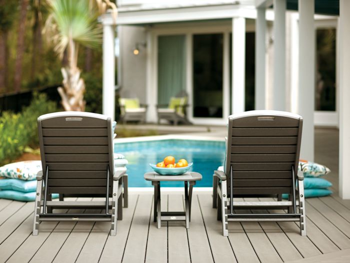 Two dark gray lounge chairs are positioned on a wooden deck facing a swimming pool, with a small table holding a bowl of oranges between them. The setting is outdoors, adjacent to a contemporary house with large glass doors and lush greenery. The scene is calm and inviting, with accent pillows stacked nearby and soft natural lighting. No people are present, emphasizing relaxation and leisure.