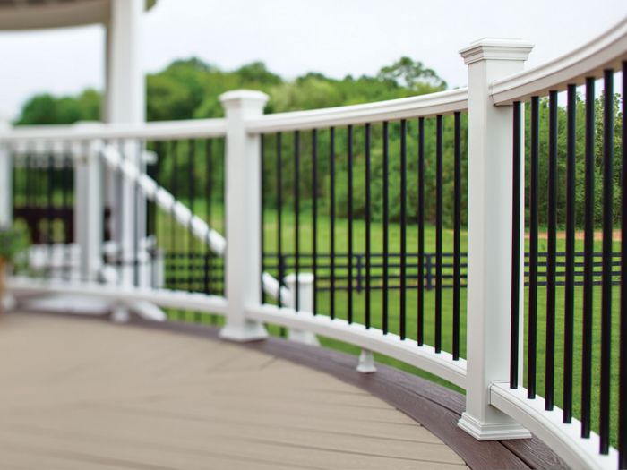 A close-up view of a curved deck railing featuring white posts and black balusters. The deck surface is a light brown composite material, and the background shows a lush green lawn with trees and a wooden fence. The setting appears to be a residential outdoor patio or porch area, with a potted plant visible on the left.