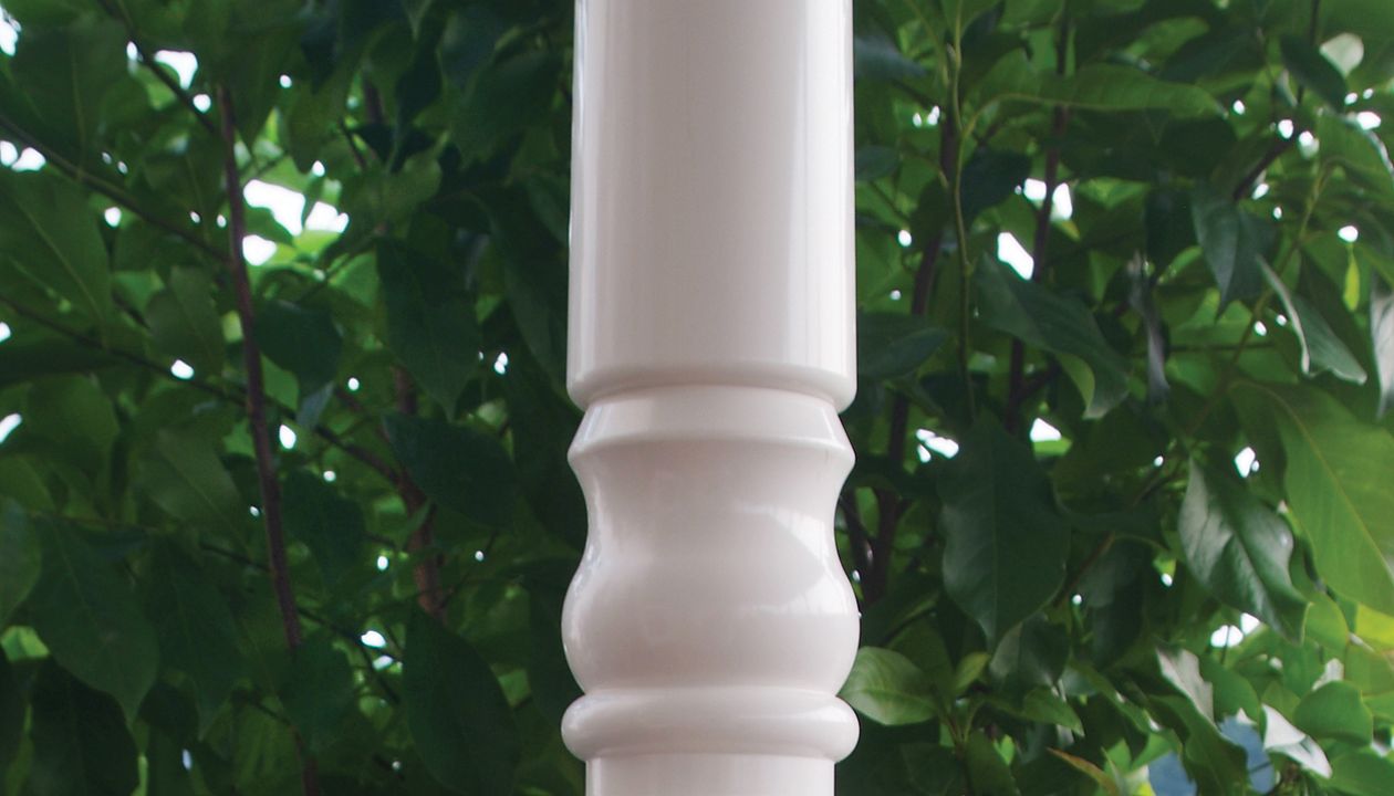 A white decorative deck post features a small round light fixture mounted on its surface. The post is part of a railing system with dark vertical balusters and a brown horizontal rail. Lush green foliage fills the background, suggesting an outdoor garden or patio setting. The image highlights the contrast between the clean architectural lines and the natural environment.