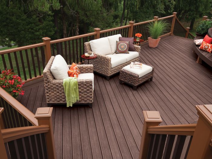 A spacious outdoor deck features brown composite flooring and comfortable wicker furniture with cream cushions. The setting includes a loveseat, armchair, ottoman, and side tables, accented by colorful pillows and potted plants. Surrounded by wooden railings and lush greenery, the area exudes a cozy and inviting atmosphere. Decorative touches like a green throw and patterned pillows add visual interest.