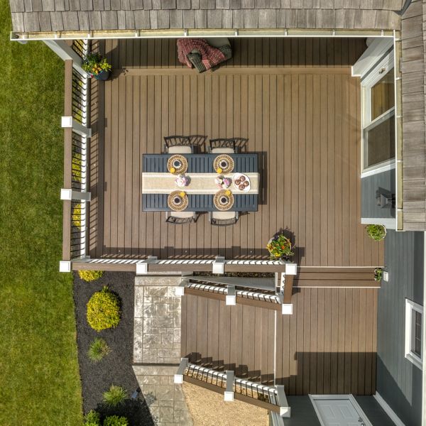 This image shows an overhead perspective of a wooden patio attached to a house. The patio features a rectangular dining table set for six, with plates and cups arranged neatly on a striped table runner. The setting is bright and inviting, with potted plants adding color and a manicured lawn visible nearby. The overall mood is clean and welcoming, ideal for outdoor gatherings.