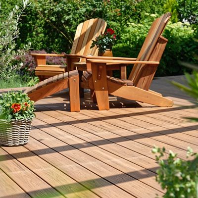 A sunlit wooden deck features two classic Adirondack chairs arranged for relaxation. The setting is enhanced by lush greenery and vibrant potted flowers, creating a tranquil outdoor retreat. Shadows from overhead beams add visual interest to the scene. The overall mood is peaceful and inviting, perfect for enjoying a quiet afternoon outdoors.