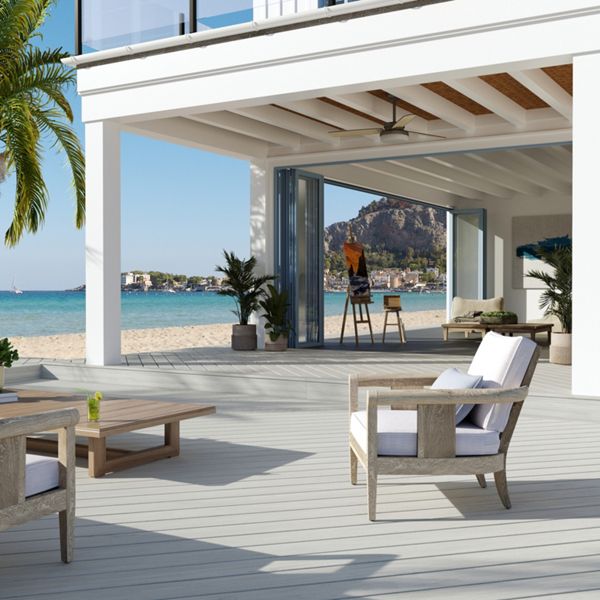 A spacious, modern patio overlooking a serene beachfront setting. The area features wooden lounge chairs with white cushions, a palm tree, and a view of the ocean and distant cliffs. The open design includes a bar area with hanging lights, creating a relaxed and inviting atmosphere.