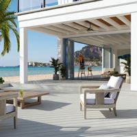 A spacious, modern patio overlooking a serene beachfront setting. The area features wooden lounge chairs with white cushions, a palm tree, and a view of the ocean and distant cliffs. The open design includes a bar area with hanging lights, creating a relaxed and inviting atmosphere.