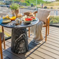 A round table set for breakfast on a balcony overlooking a serene ocean view. The table features fresh fruit, orange juice, bread, and cereal, accompanied by cozy seating with cushions and a blanket. The setting is bright and inviting, with clear skies and a calm waterfront in the background.