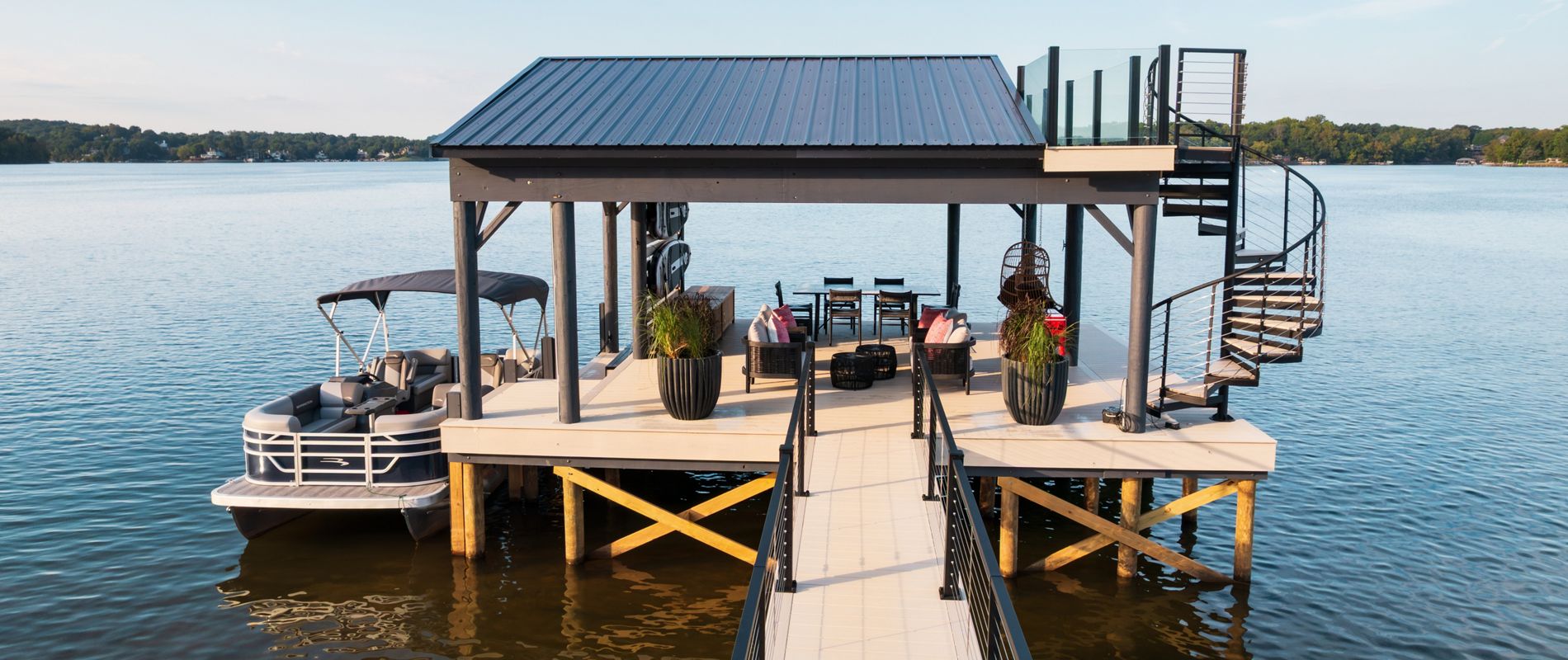 A Trex Transend Lineage dock in the color Salt Flat on Lake Wiley. A boat is docked and there is a lovely sitting area underneath a roofed covering and the dock features a Trex Spiral Staircase leading to the second floor for more views of the lake.