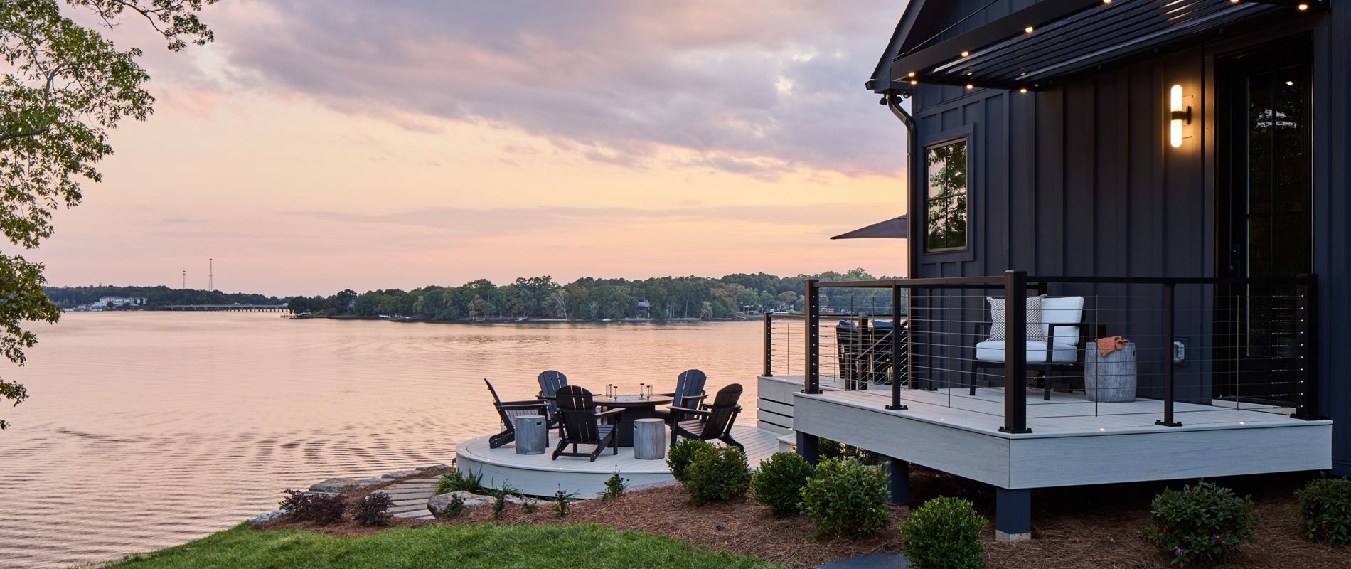 A view of the house and the Trex Transcend Lineage deck in Salt Flat, Trex Signature X-Series Cable Railing, and Trex outdoor furniture and lighting options overlooking Lake Wiley as the sun sets, casting beautiful colors in the sky and on the water.