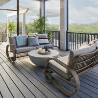 A contemporary screened-in porch features two rattan lounge chairs with plush cushions and patterned throw pillows. The setting includes a round coffee table with decorative items and potted plants, all under a ceiling fan. The space is bright and airy, with natural light, neutral tones, and views of greenery beyond the railing.