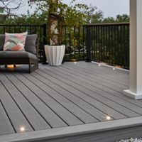 A contemporary outdoor deck features gray composite flooring, a cushioned chair with a colorful geometric pillow, and a large potted plant. The setting is enhanced by built-in deck lights and a black metal railing, with a pergola overhead and a cloudy sky in the background. The overall mood is calm and inviting, ideal for relaxation or small gatherings.