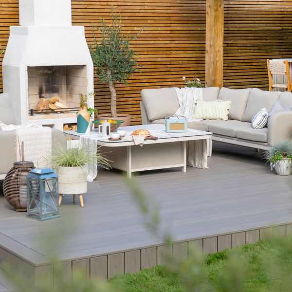 A stylish outdoor patio featuring a white fireplace surrounded by comfortable beige sofas and a coffee table. The setting includes decorative lanterns, plants, and wooden accents, creating a warm and inviting atmosphere. The background showcases a wooden panel wall and a bar area with stools.