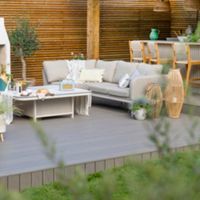 A stylish outdoor patio featuring a white fireplace surrounded by comfortable beige sofas and a coffee table. The setting includes decorative lanterns, plants, and wooden accents, creating a warm and inviting atmosphere. The background showcases a wooden panel wall and a bar area with stools.