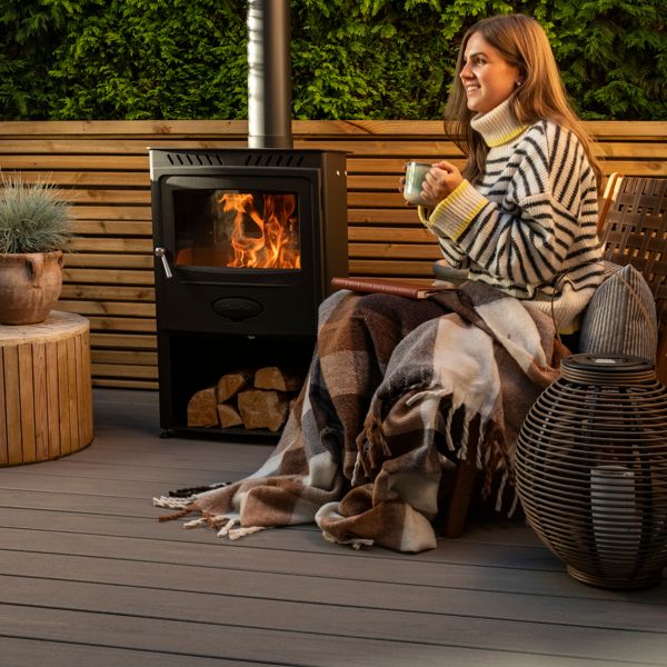 A warm outdoor setting featuring a wood-burning stove surrounded by wooden furniture and decor. The area includes potted plants, woven baskets, and a striped blanket for added comfort. The ambiance is enhanced by the glowing fire and natural wooden textures, creating a relaxing atmosphere.