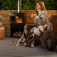 A warm outdoor setting featuring a wood-burning stove surrounded by wooden furniture and decor. The area includes potted plants, woven baskets, and a striped blanket for added comfort. The ambiance is enhanced by the glowing fire and natural wooden textures, creating a relaxing atmosphere.