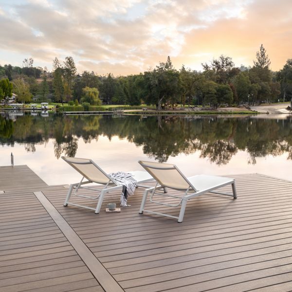 Two modern white lounge chairs sit on a spacious wooden dock overlooking a calm lake. The tranquil setting features a soft sunrise sky, gentle reflections on the water, and lush trees along the shoreline. A striped towel and a cup rest on one of the chairs, suggesting relaxation and leisure. No visible text or numbers are present in the image.