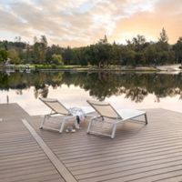 Two modern white lounge chairs sit on a spacious wooden dock overlooking a calm lake. The tranquil setting features a soft sunrise sky, gentle reflections on the water, and lush trees along the shoreline. A striped towel and a cup rest on one of the chairs, suggesting relaxation and leisure. No visible text or numbers are present in the image.