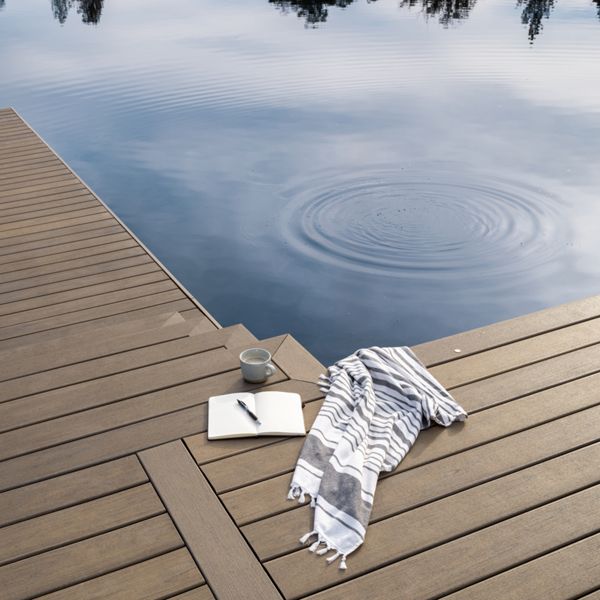 A serene lakeside scene features a wooden dock extending over still water, with a notebook, pen, cup, and striped towel arranged on the planks. Gentle ripples are visible on the lake’s surface, reflecting the sky and trees. The setting suggests a peaceful moment for writing or relaxation in a natural outdoor environment.