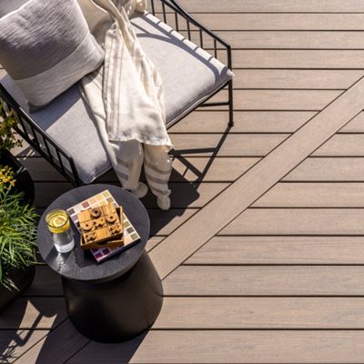 A contemporary outdoor lounge chair with plush cushions and a draped throw sits on a sunlit wooden deck. Beside the chair, a small round table holds a candle, a wooden puzzle, and a magazine, adding a cozy and inviting touch. The scene is bright and tranquil, with neutral tones and clean lines. No people are present, emphasizing relaxation and leisure.