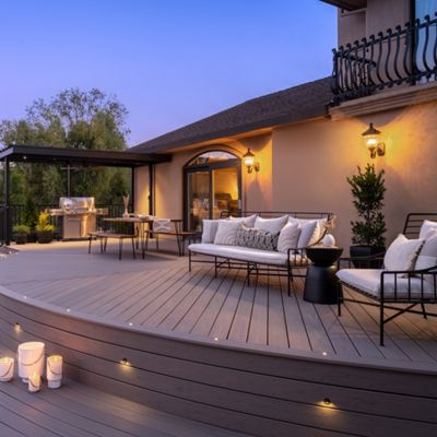 A spacious outdoor patio featuring modern furniture and soft lighting. The setting includes a curved wooden deck adorned with candles and a dining area under a pergola. The tranquil evening ambiance is complemented by lush greenery and a serene backdrop.