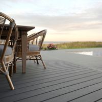 A spacious outdoor deck features a contemporary dining set with wicker chairs and a wooden table. The scene overlooks a serene swimming pool surrounded by lounge chairs and lush landscaping. The setting is tranquil, with soft natural lighting and a neutral color palette, evoking a relaxed, inviting mood.